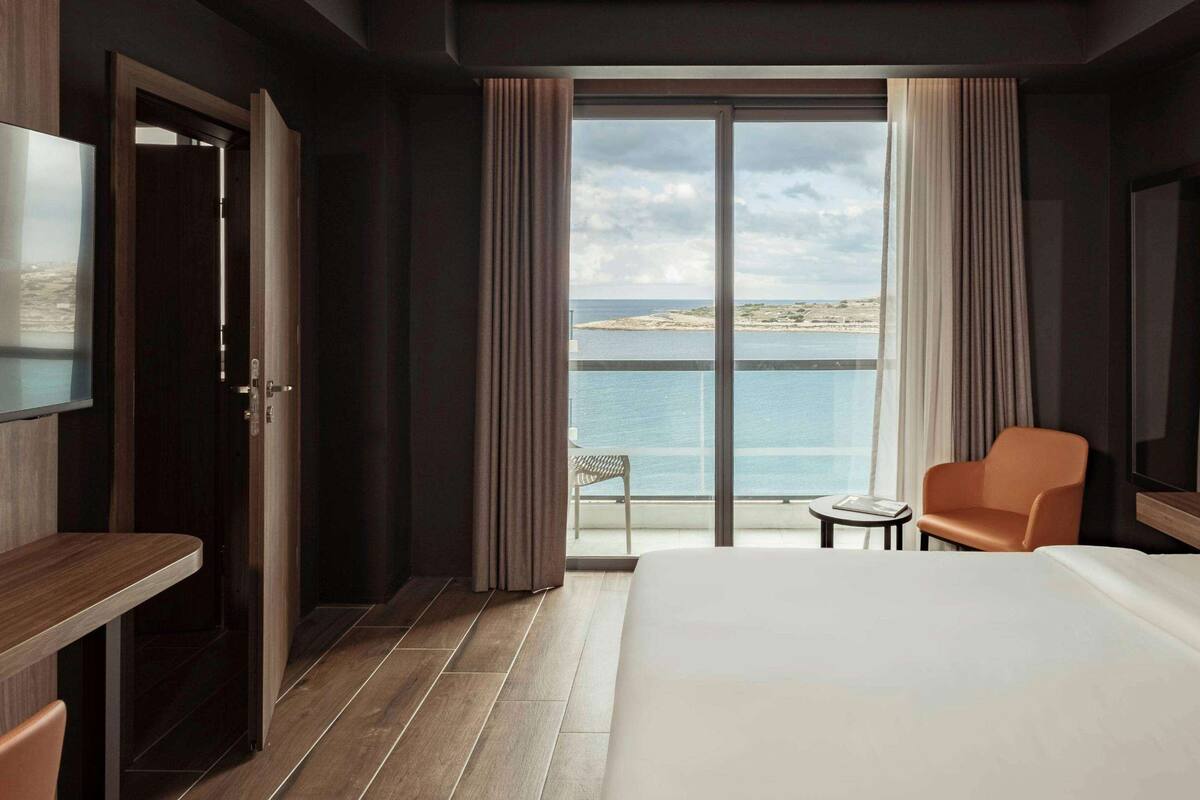 A hotel room with a large bed, a desk, a chair, and a small table. There is a large window with a view of the sea and a distant island.