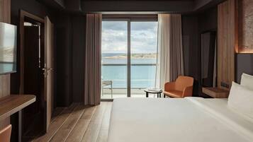 Une chambre d’hôtel avec un grand lit, un bureau, une chaise et une petite table. Une grande fenêtre offre une vue sur la mer et une île lointaine.