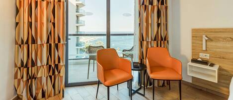A modern hotel room with a balcony, two orange chairs, a small table, and a wooden headboard.