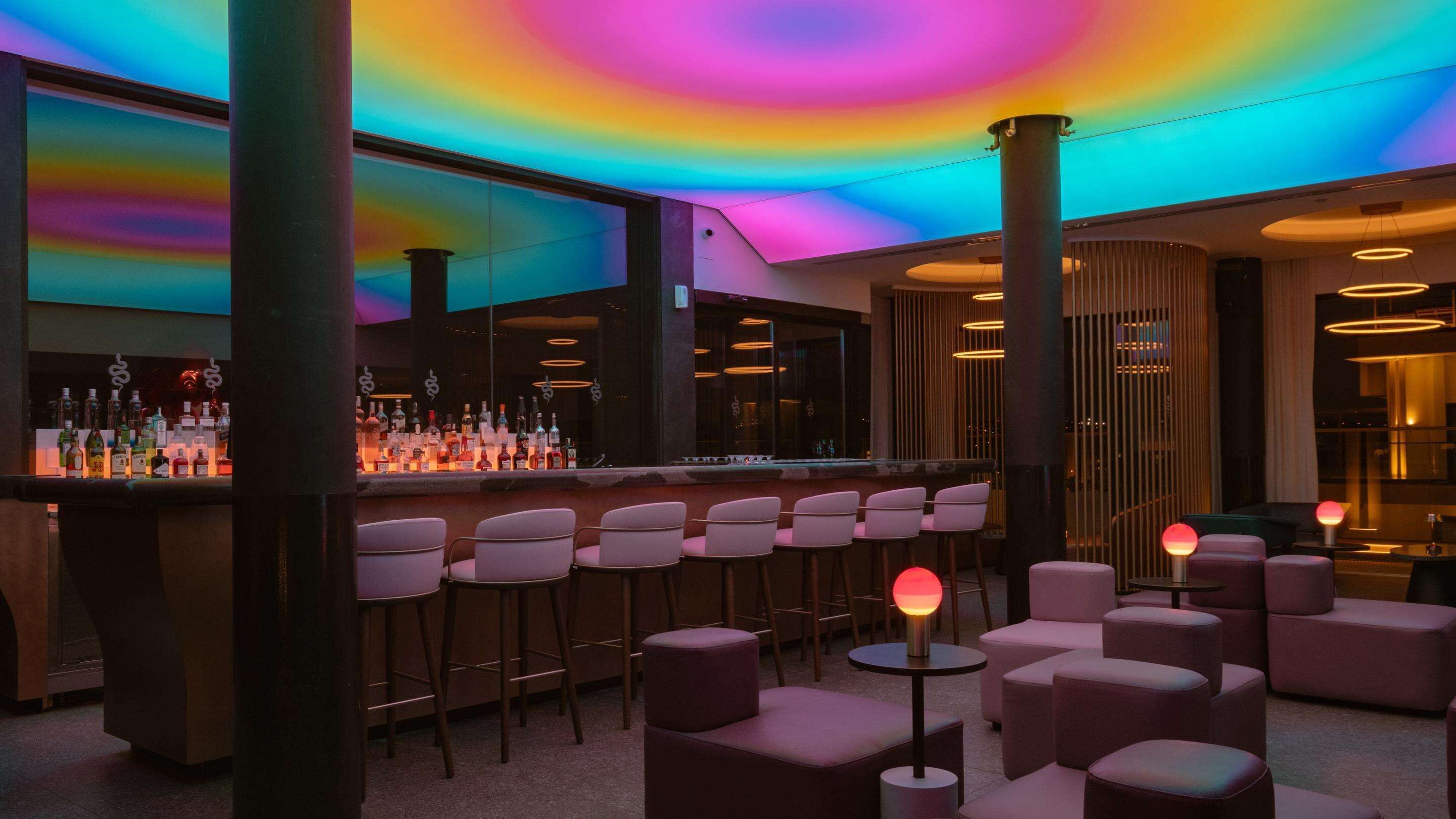 A bar area with a colorful ceiling, a bar counter with various bottles, and several seating options.