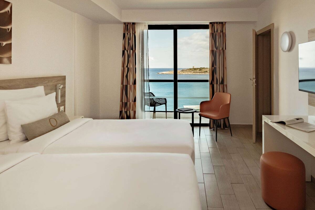 A hotel room with two beds, a desk, a chair, and a large window offering a view of the sea.