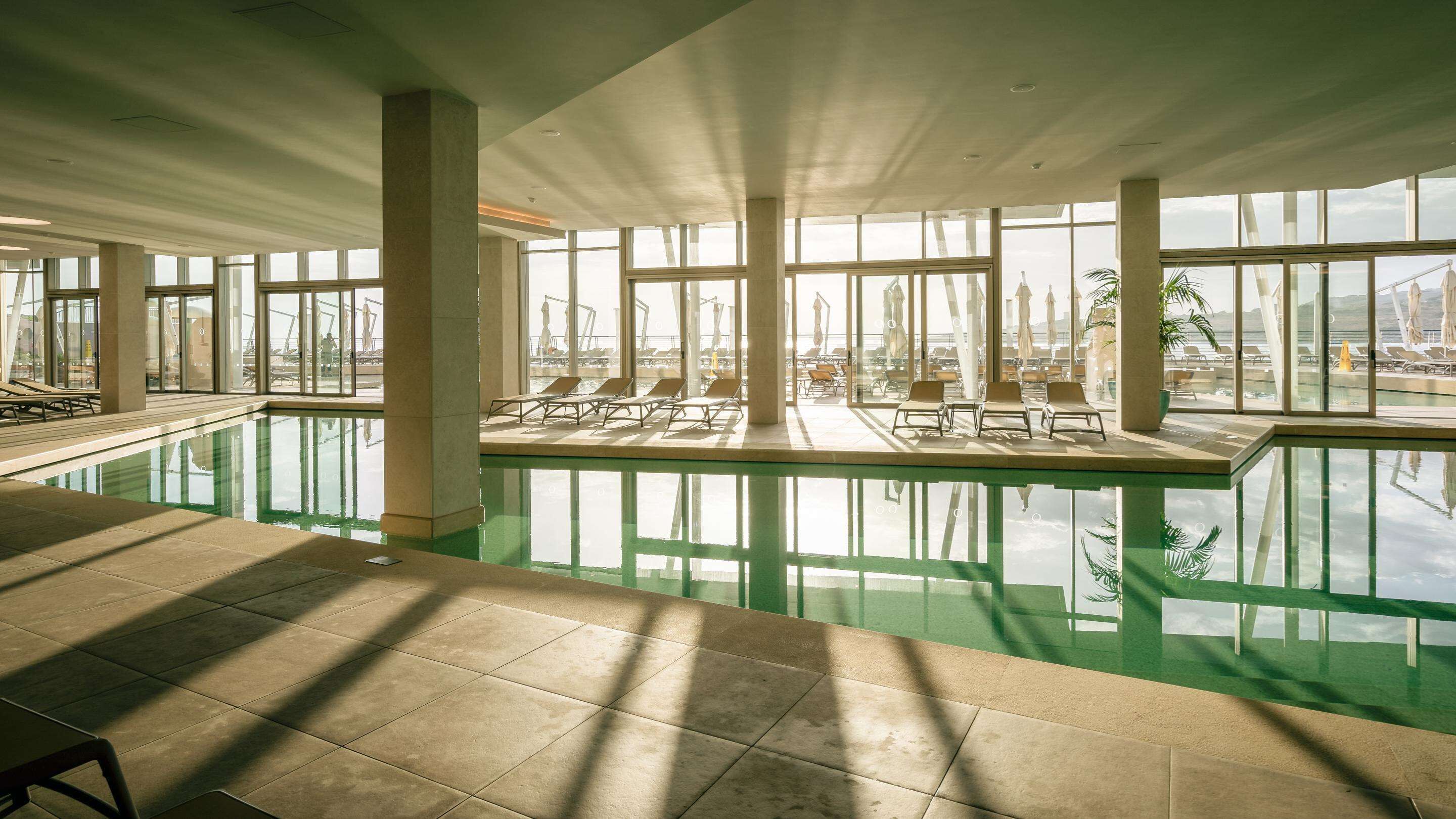 Una piscina interna con vista aperta sull'esterno, interni luminosi e sedie a sdraio.