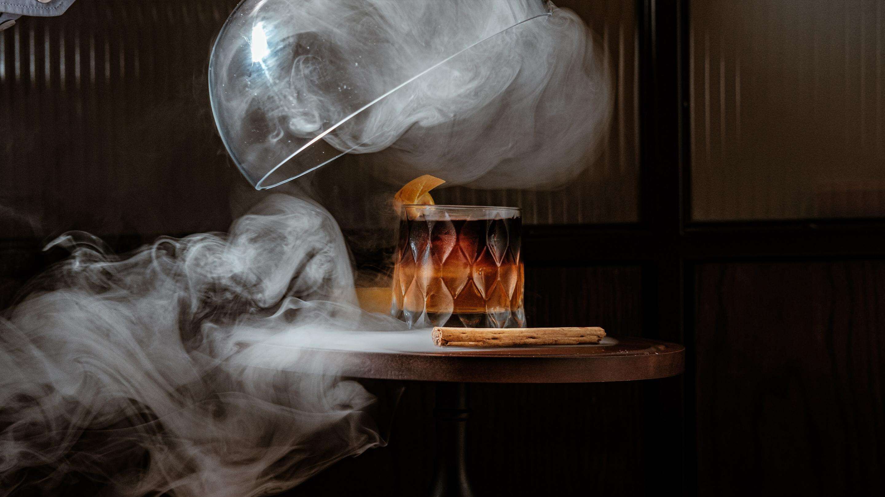 Un verre d’une boisson de couleur ambrée, agrémenté d’une garniture et d’une tige de cannelle, ainsi qu’un verre d’eau, posés sur une table en bois.