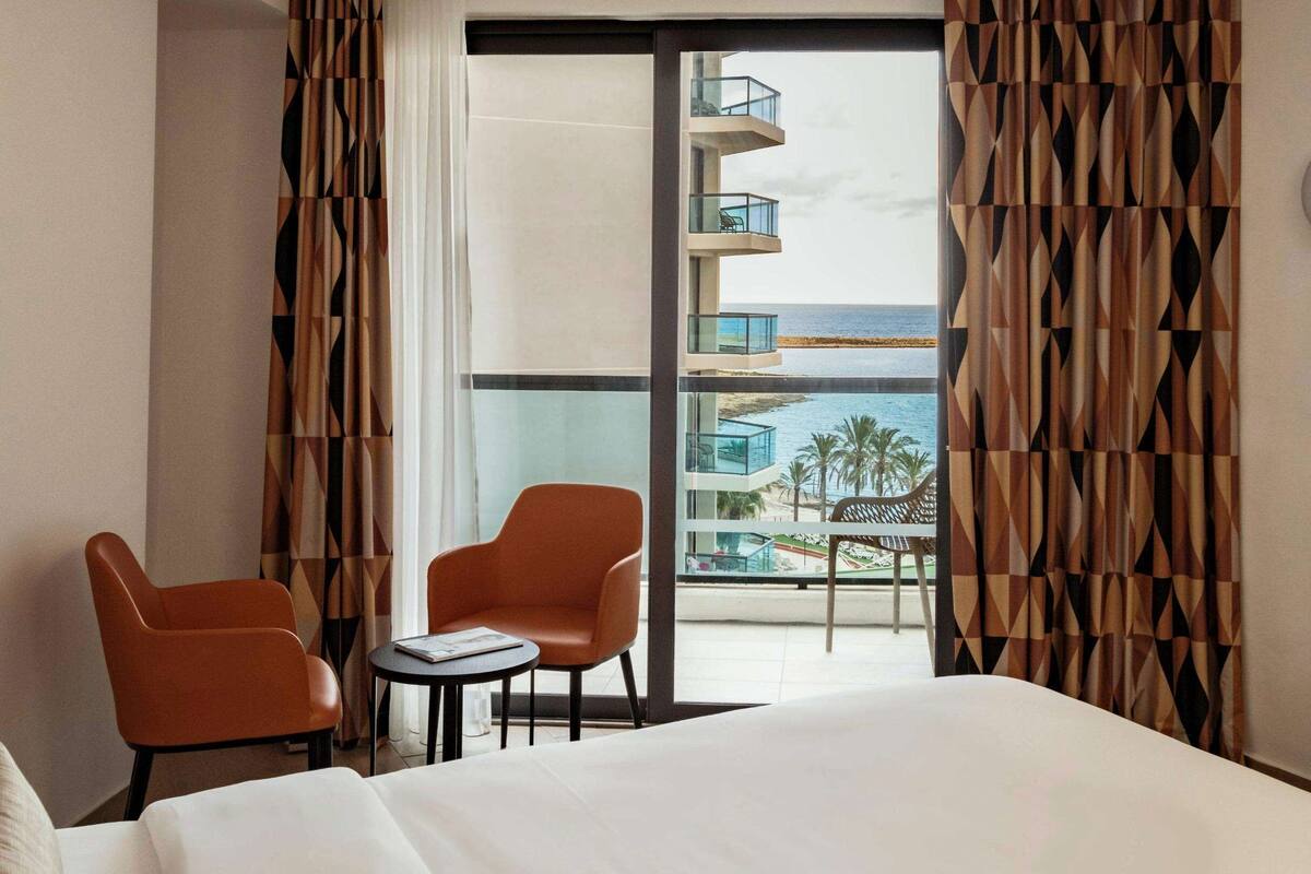 A hotel room with a bed, two chairs, a small table, and a view of a beach and ocean through large windows.