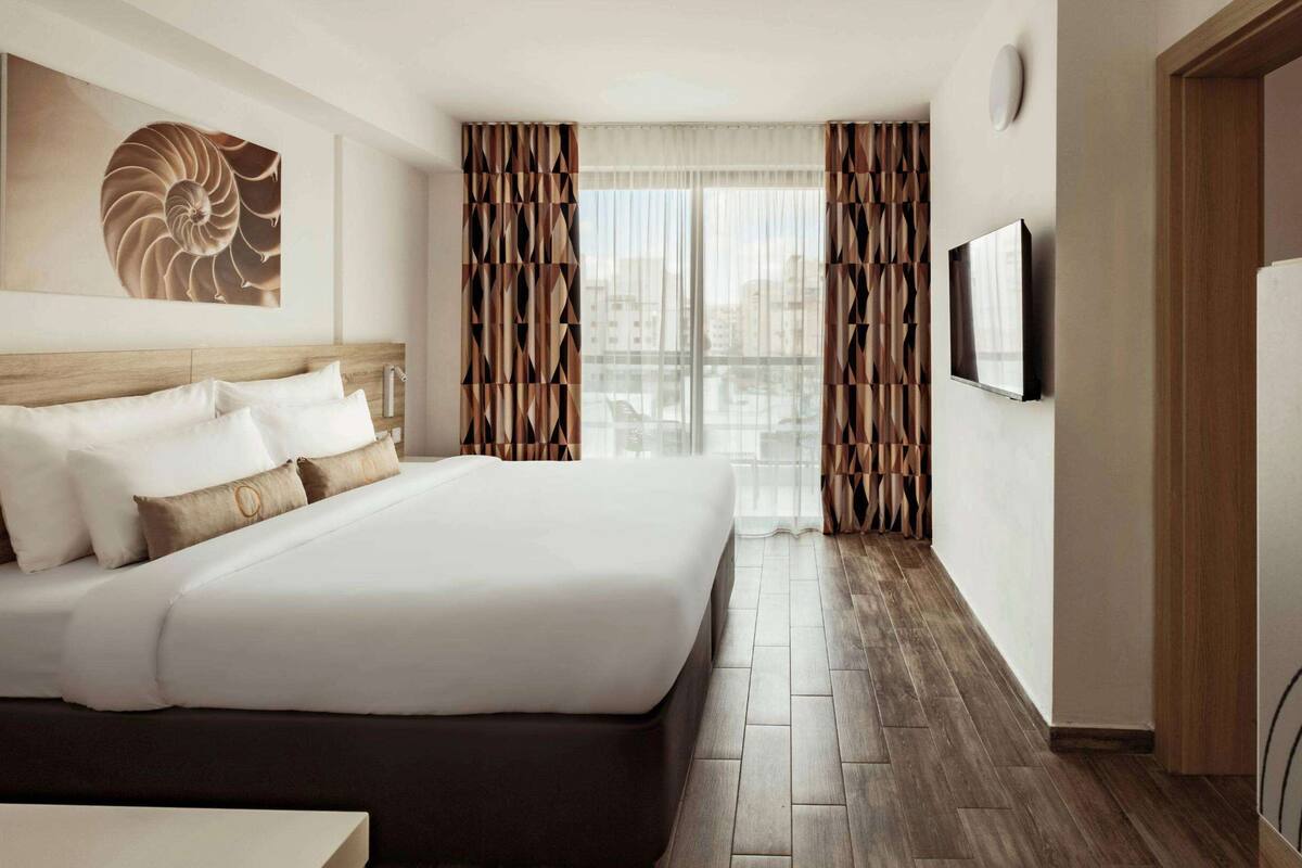A hotel room with a large bed, a wooden headboard, a wall-mounted TV, and a city view through the window.
