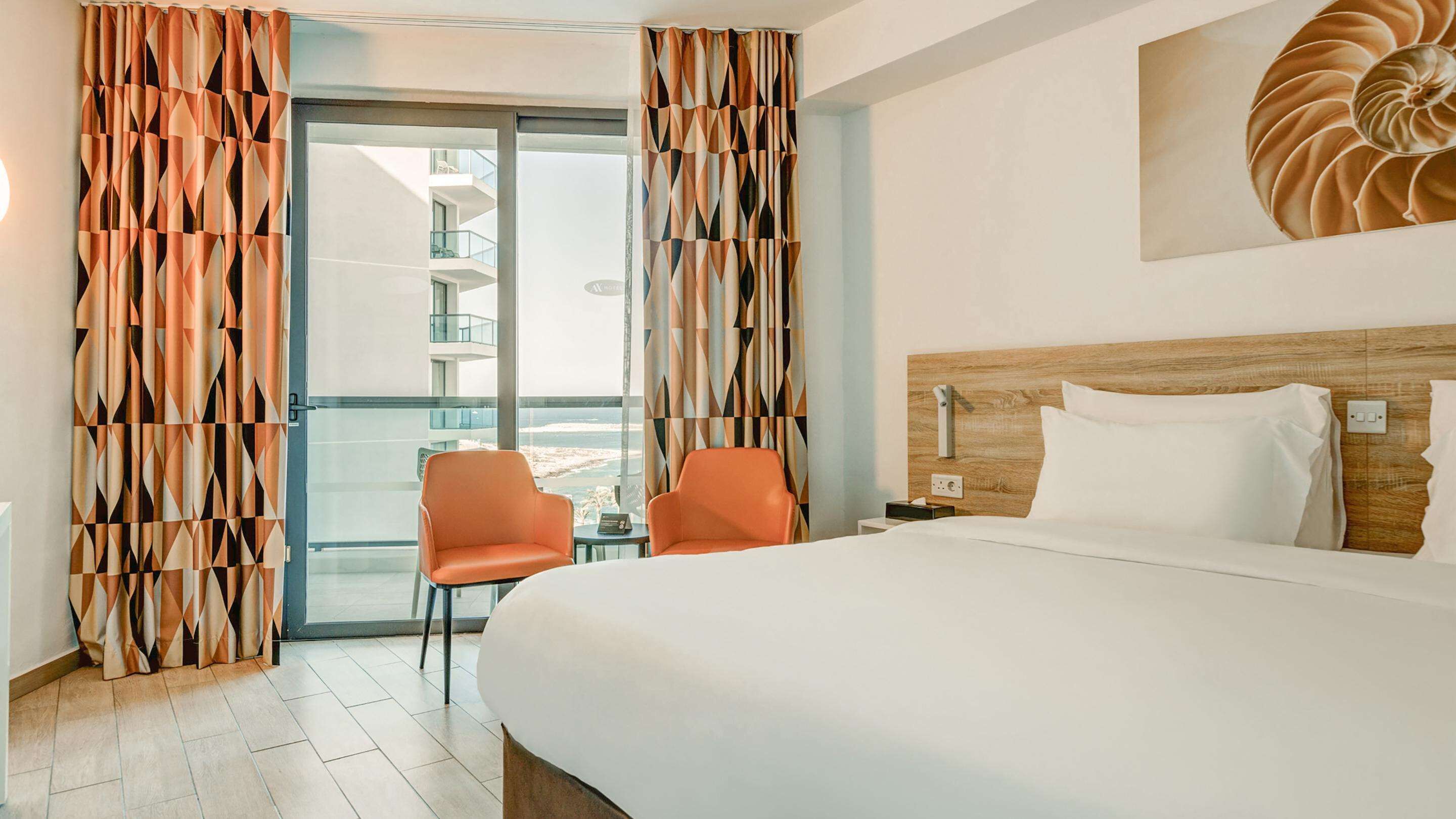 Een hotelkamer met een groot bed, twee oranje stoelen, een houten hoofdeinde, uitzicht op een gebouw door een glazen deur en een kunstwerk aan de muur.