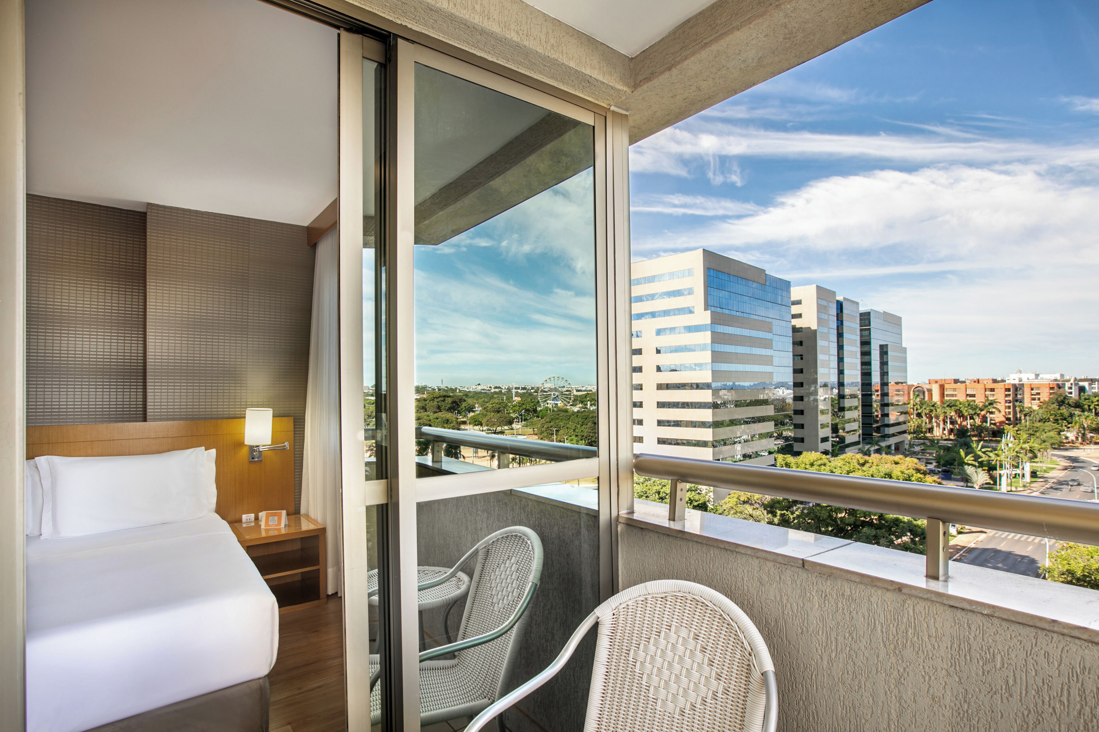 A hotel room with a bed, a nightstand, a lamp, a chair, and a balcony with a view of buildings and greenery.