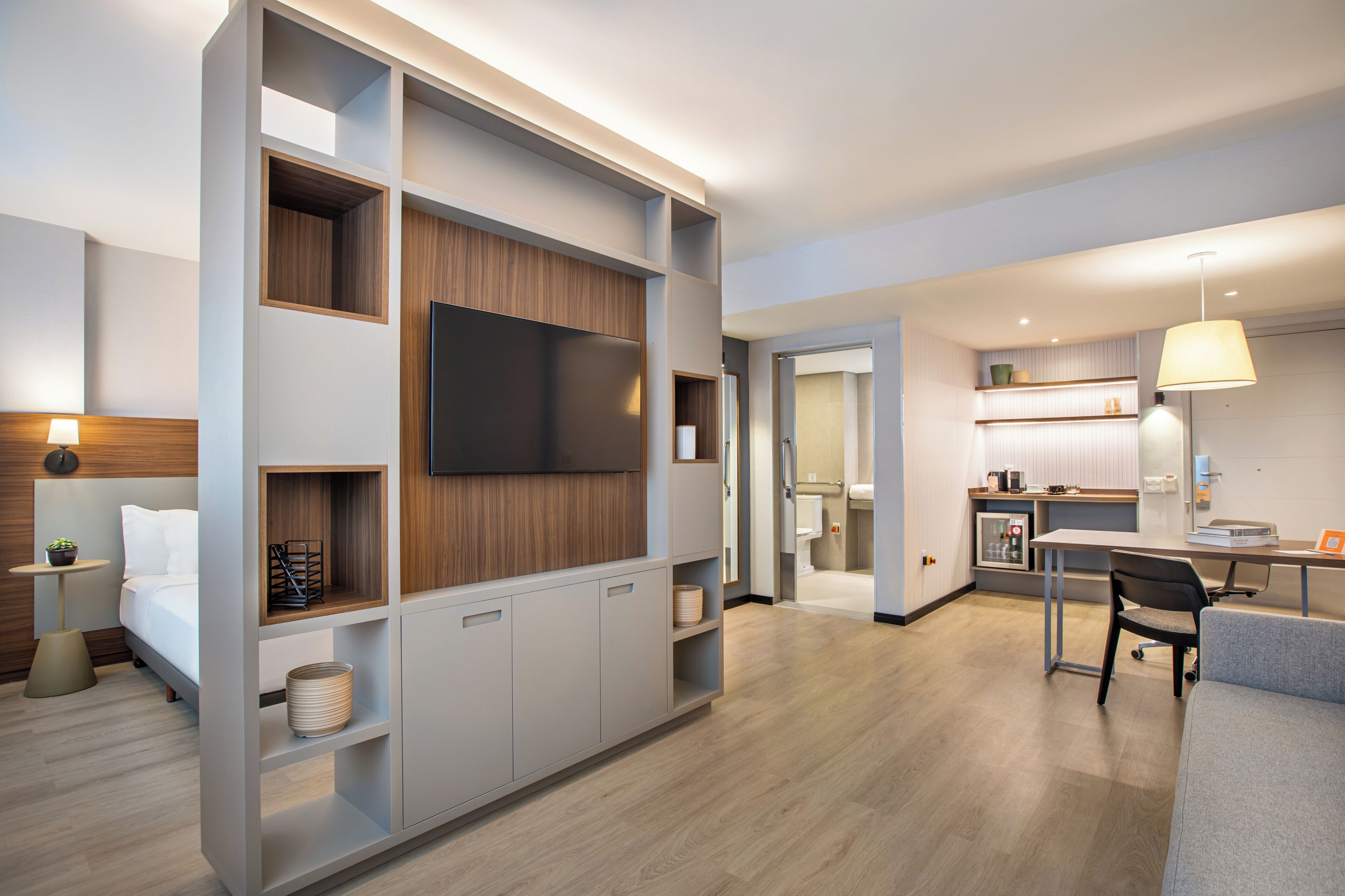 Una habitación de hotel moderna con una cama, un televisor montado en la pared, un escritorio con una silla y una zona de kitchenette.