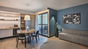 Een moderne woonkamer met een bank, een eethoek met stoelen en een tafel, een keuken met kastjes en een koffieapparaat, en een groot raam met uitzicht.