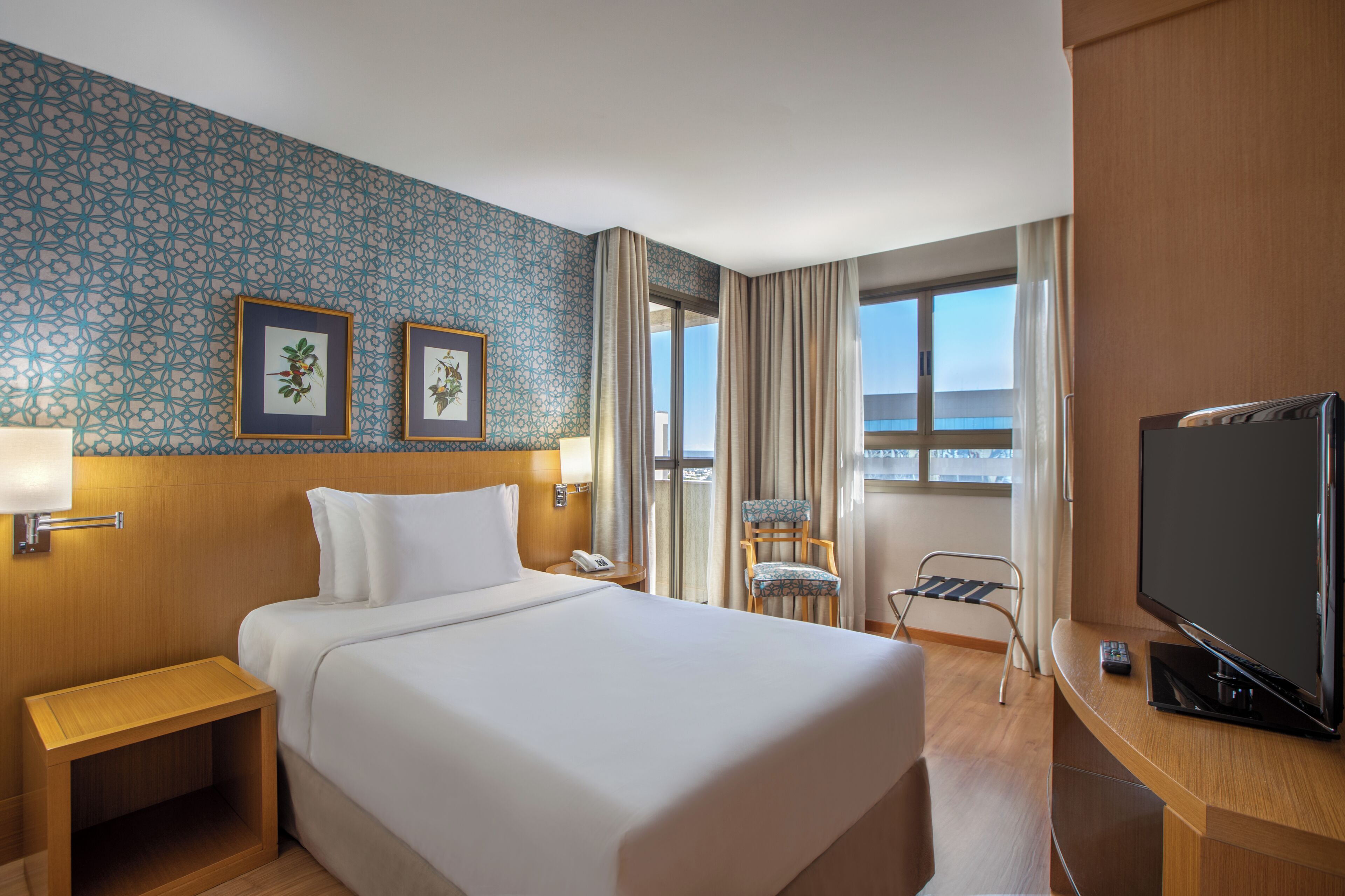Quarto de hotel com uma cama grande, televisão, escrivaninha, cadeira, janela com cortinas e duas imagens emolduradas na parede.