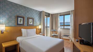 Quarto de hotel com uma cama grande, televisão, escrivaninha, cadeira, janela com cortinas e duas imagens emolduradas na parede.