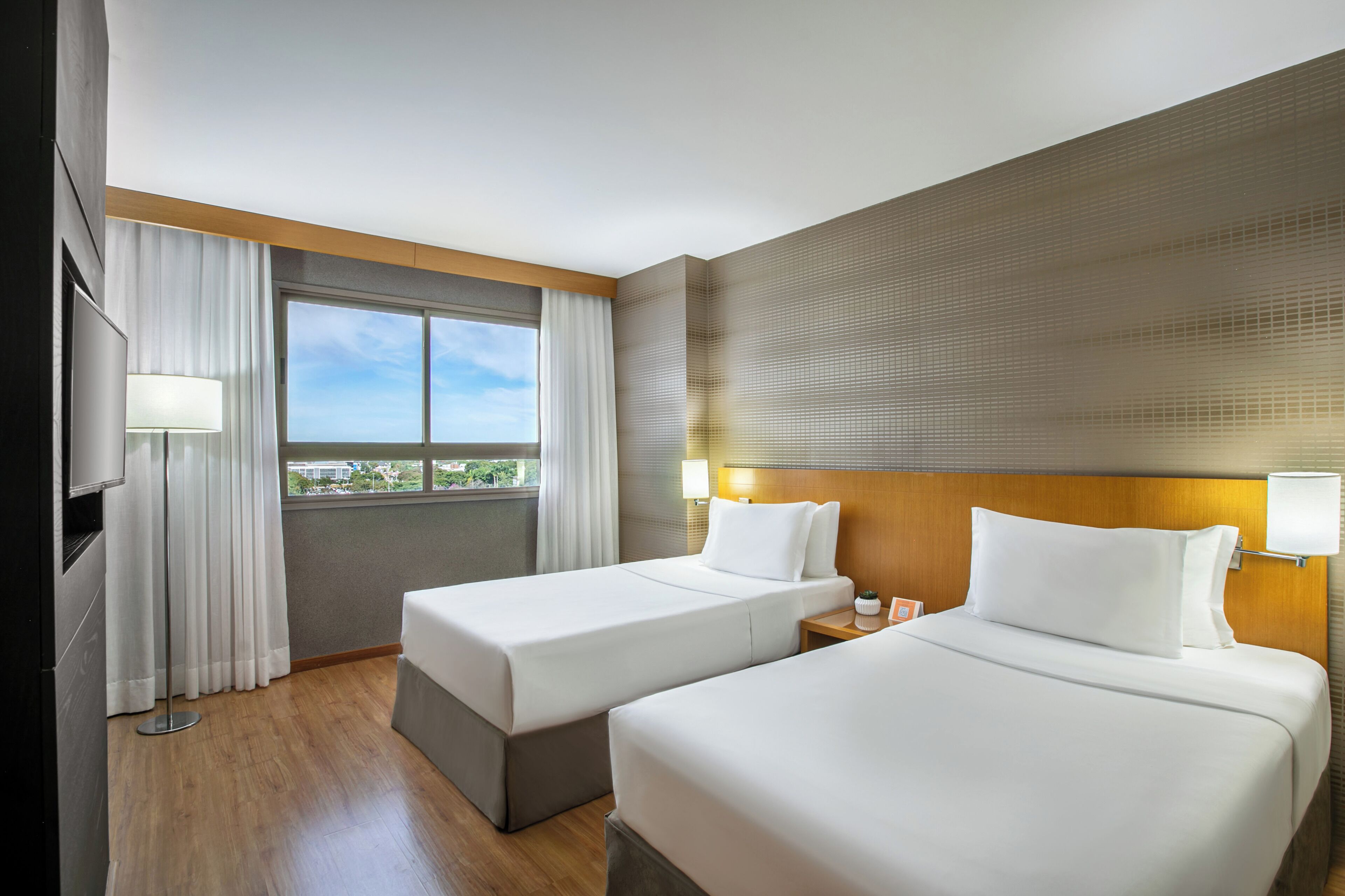 A hotel room with two beds, a TV, a window with curtains, and a view of buildings outside.