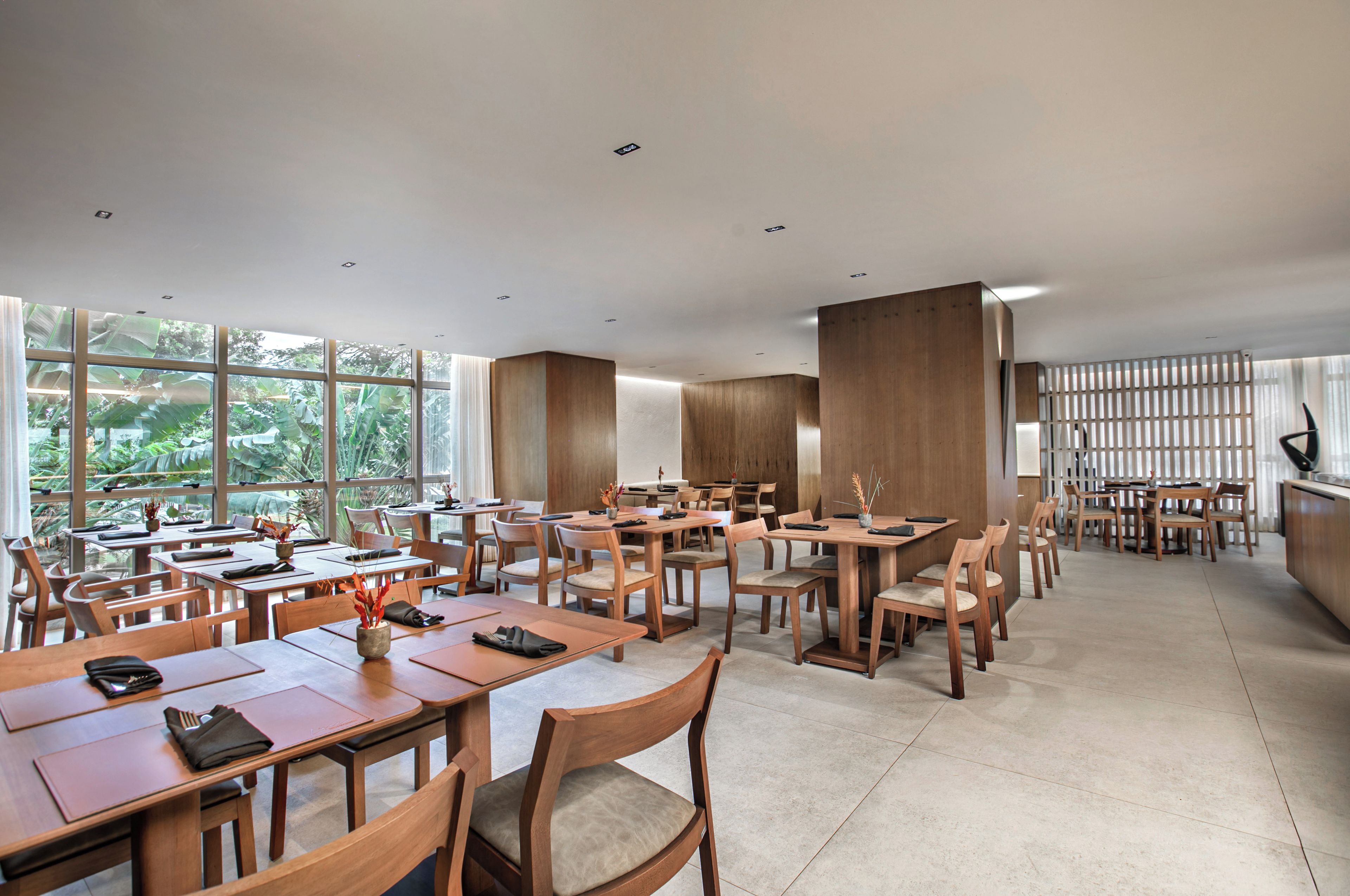 A spacious dining area with multiple tables and chairs, large windows offering a view of greenery, and a modern interior design.