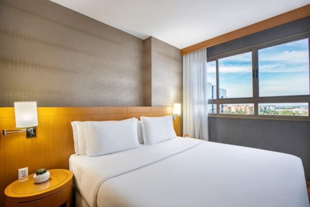 Une chambre d’hôtel avec un grand lit, une table de chevet, une lampe et une fenêtre donnant sur la ville.