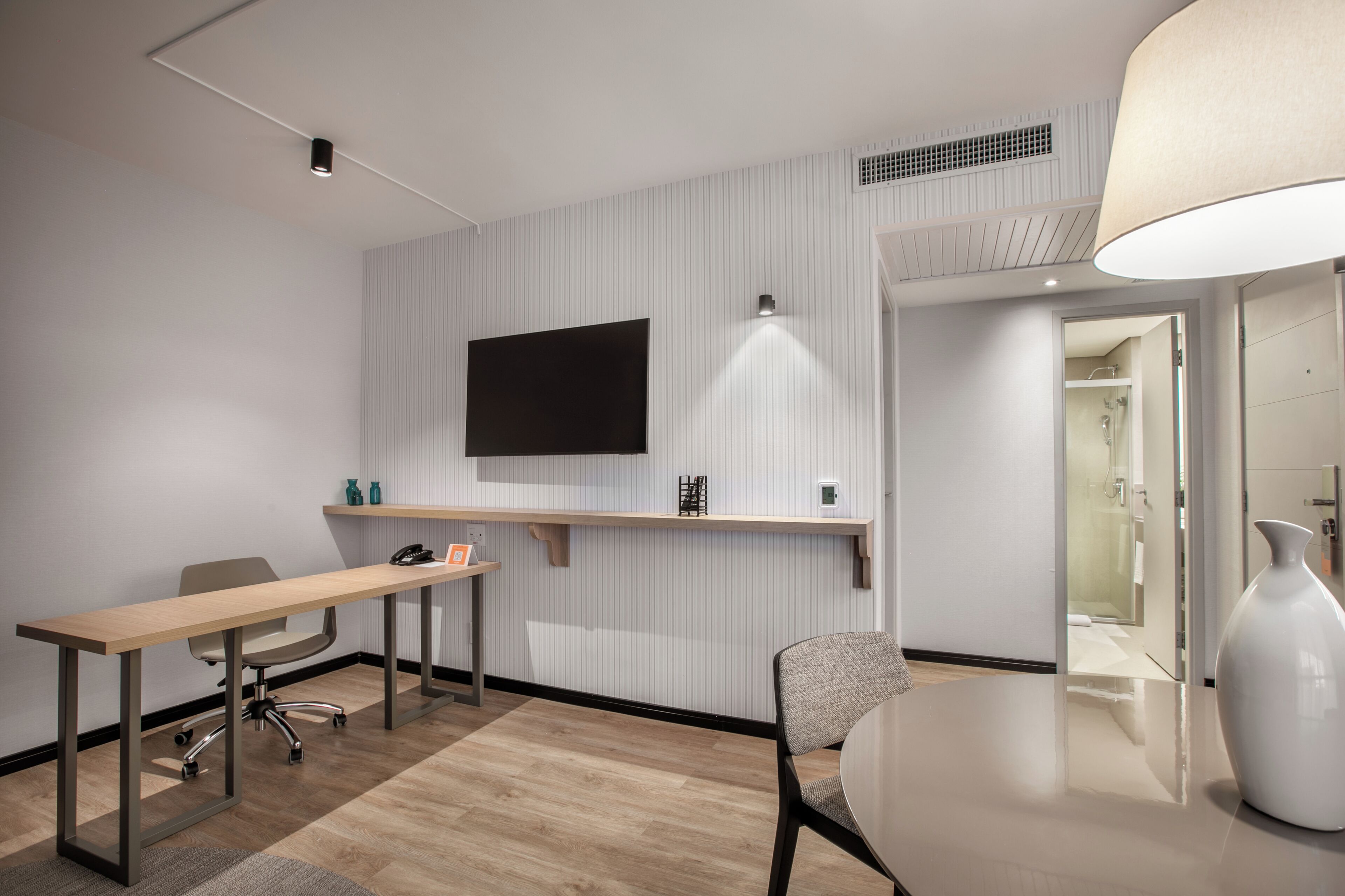 Una habitación de hotel moderna con un escritorio de madera, una silla, un televisor montado en la pared y un jarrón grande sobre la mesa.