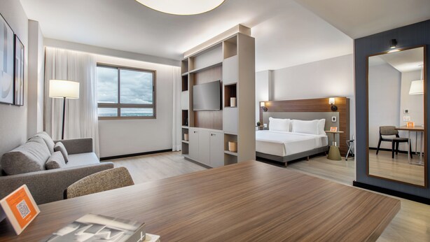 A modern hotel room with a large bed, a seating area with a sofa and chairs, a dining table, and a built-in TV shelf.