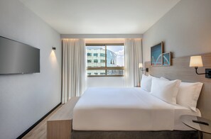 A hotel room with a large bed, a flat-screen TV, a window with curtains, a lamp, and a painting on the wall. - Brasil 21 Suites Affiliated by Melia (Brasília)