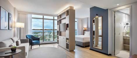 A modern hotel room with a large window, a bed, a seating area with a sofa and armchair, a small table, and a built-in bookshelf.