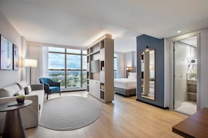 Una habitación de hotel moderna con un ventanal grande, una cama, una zona de estar con sofá y sillón, una mesita y una estantería empotrada.