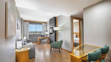 A hotel room with a sofa, a small table, a glass-top dining table, a chair, a lamp, and a view of the city.