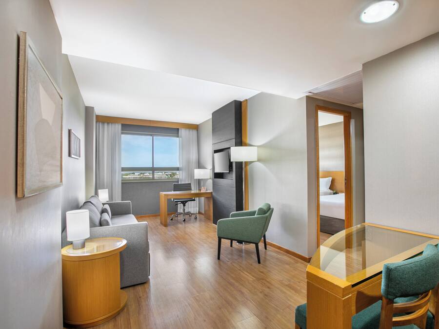 A hotel room with a sofa, a small table, a glass-top dining table, a chair, a lamp, and a view of the city.