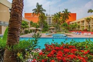 Zona de piscina de hotel con palmeras, un puente y sillones de descanso.