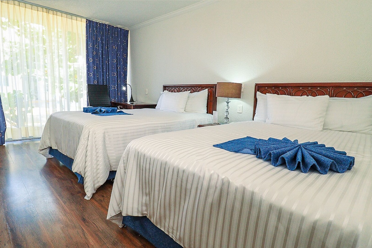 Habitación de hotel con dos camas, cabeceros de madera, ropa blanca, televisor y una ventana con cortinas azules.
