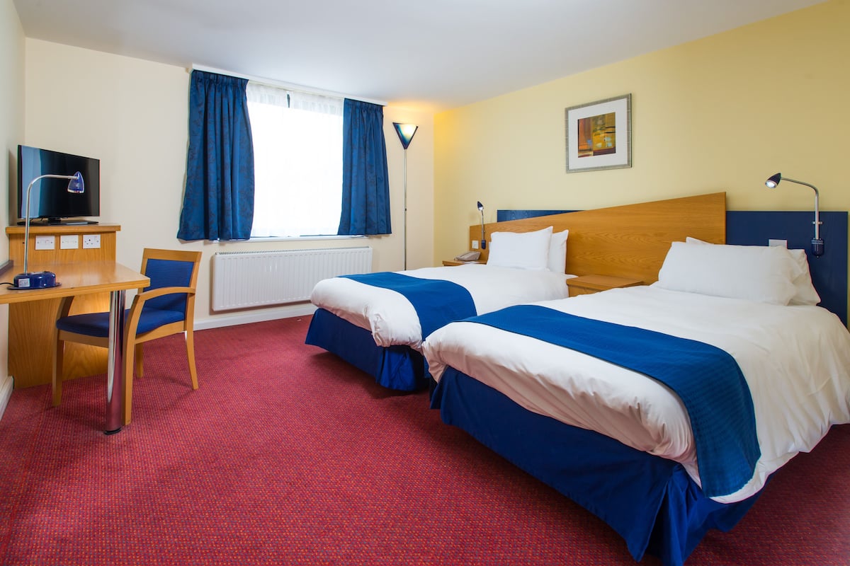 A hotel room with two beds, a desk, a chair, a TV, and a window with curtains.