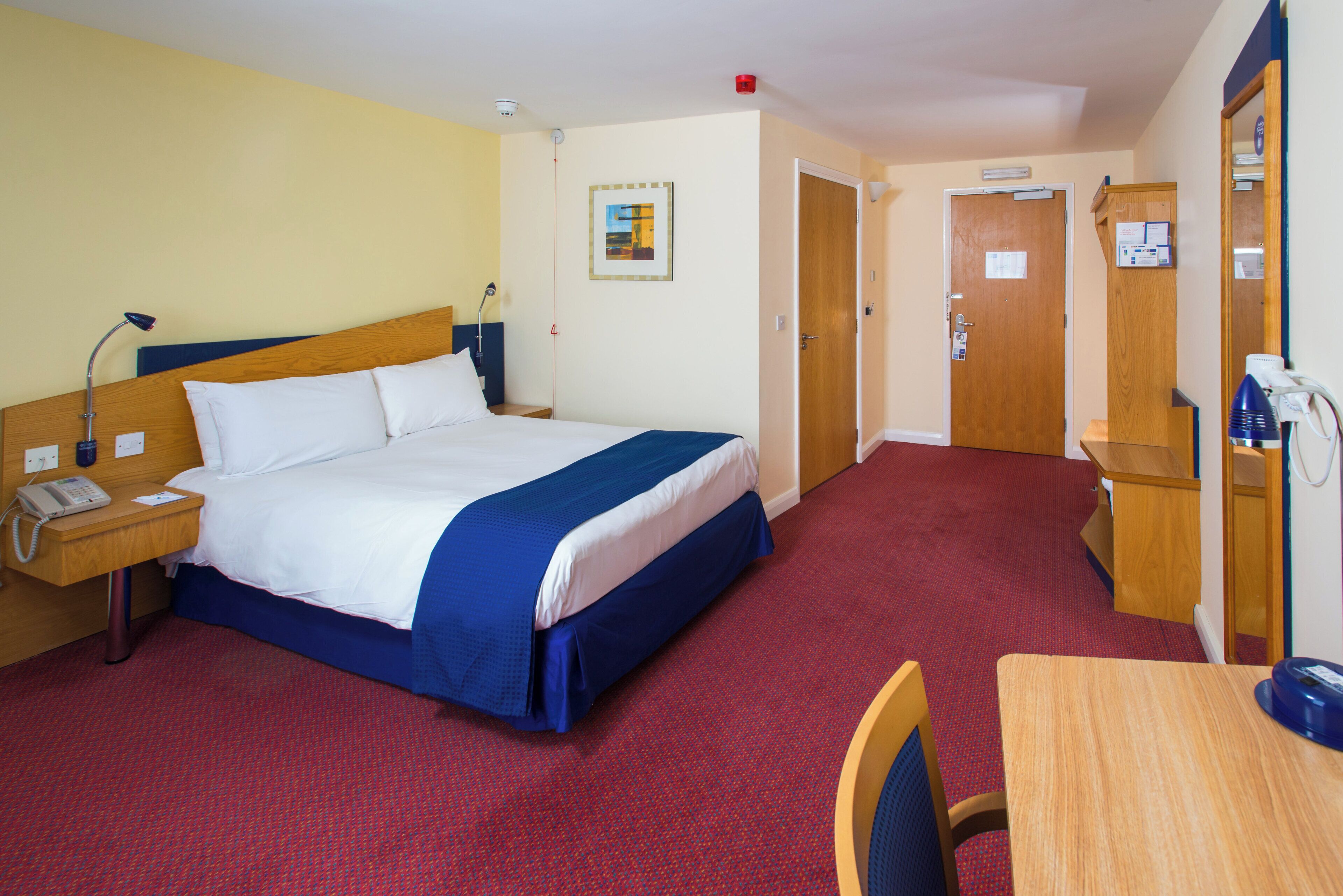 A hotel room with a bed, bedside table, lamp, desk, chair, and a red carpeted floor.