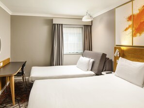 A hotel room with two beds, a desk, a chair, a window with curtains, and a wall-mounted light fixture. - ibis Styles Birmingham Centre (Birmingham)