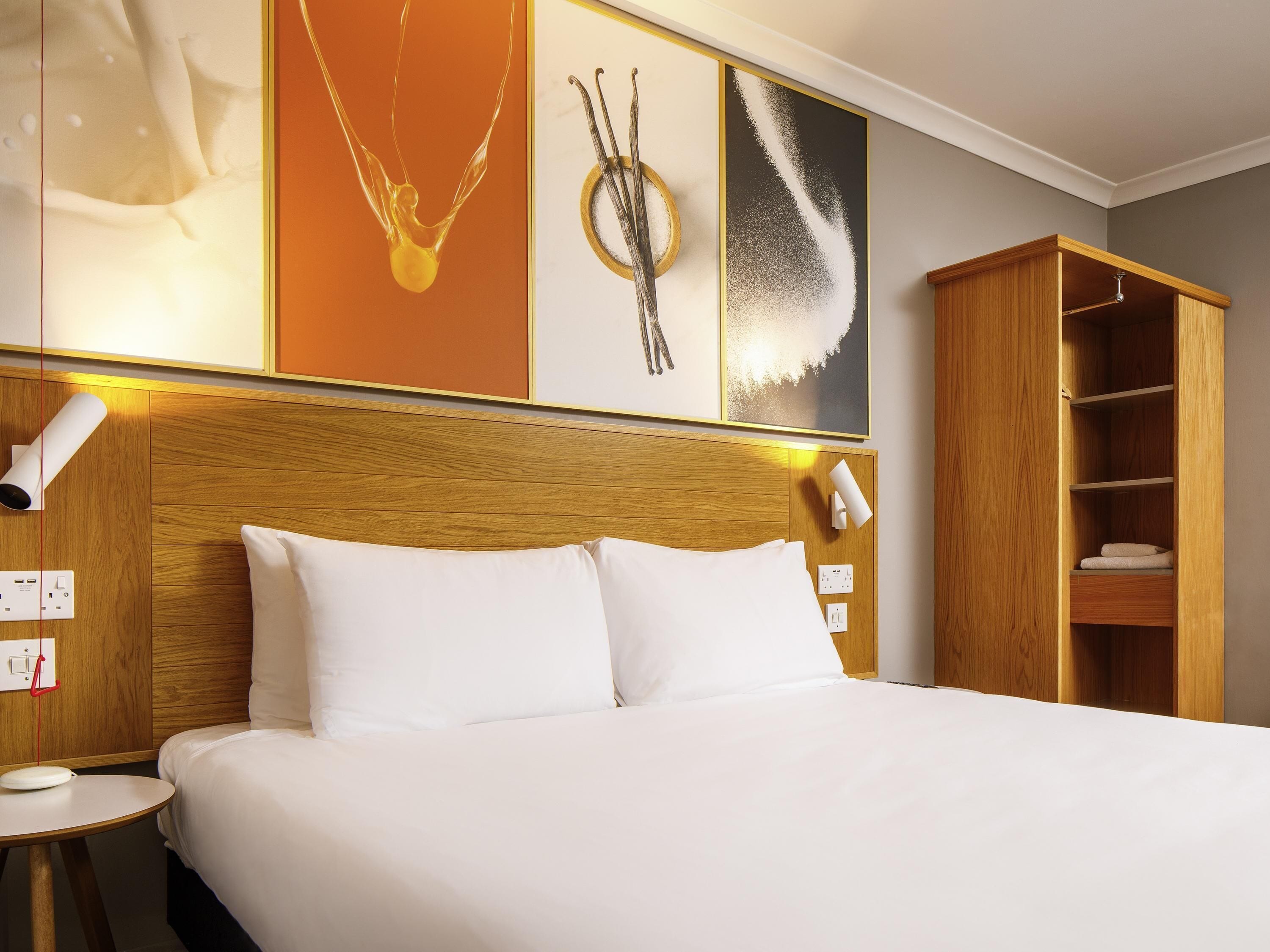 A hotel room with a wooden headboard, white bedding, a bedside table with a lamp, and a wardrobe with shelves.