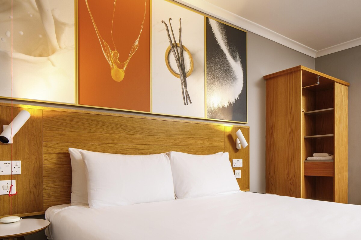 A hotel room with a wooden headboard, white bedding, a bedside table with a lamp, and a wardrobe with shelves.