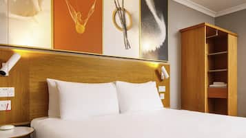 A hotel room with a wooden headboard, white bedding, a bedside table with a lamp, and a wardrobe with shelves.