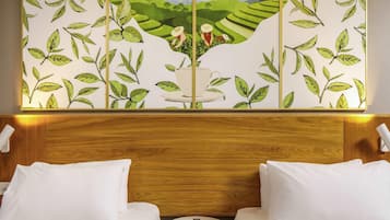 Quarto de hotel com um mural de paisagem verde na parede, duas camas brancas e uma mesa redonda pequena com um controle remoto.