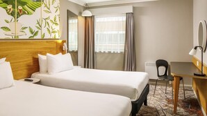 A hotel room with two beds, a desk, a chair, a large window with curtains, and a wall-mounted mirror.