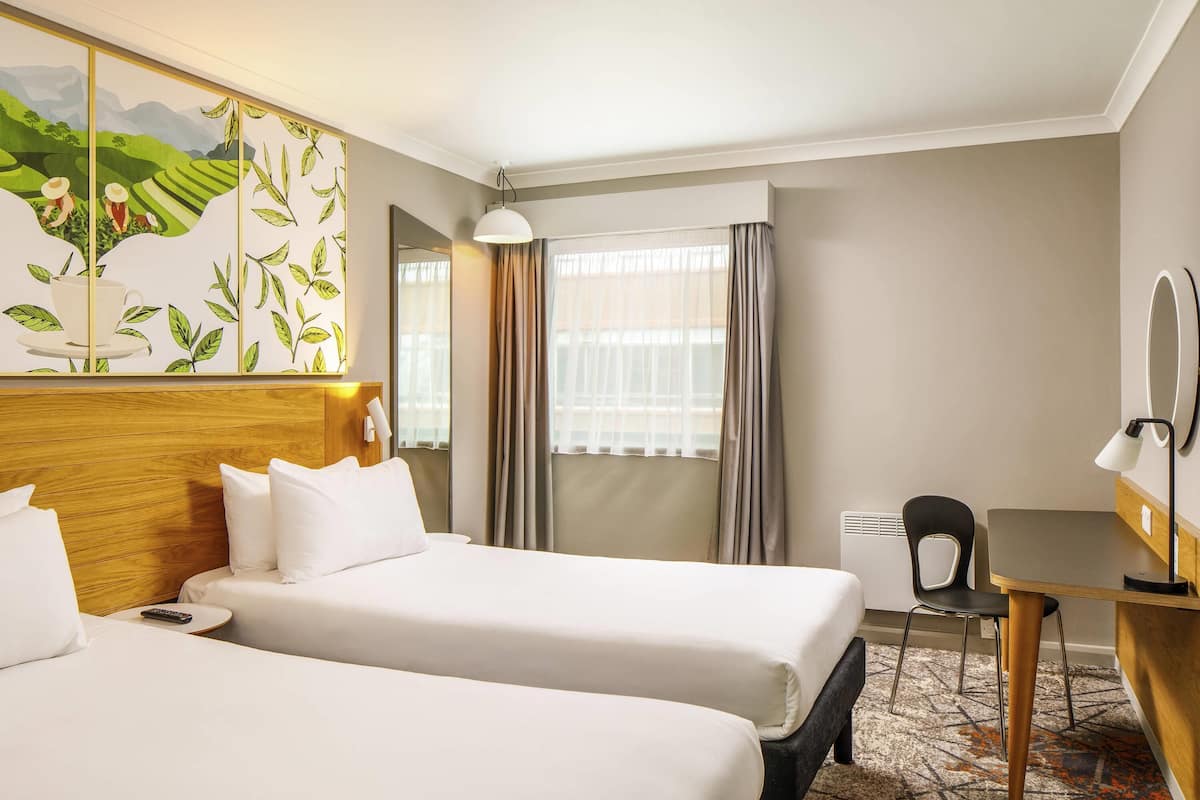 A hotel room with two beds, a desk, a chair, a lamp, a mirror, and a window with curtains.