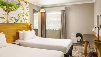 A hotel room with two beds, a desk, a chair, a lamp, a mirror, and a window with curtains.