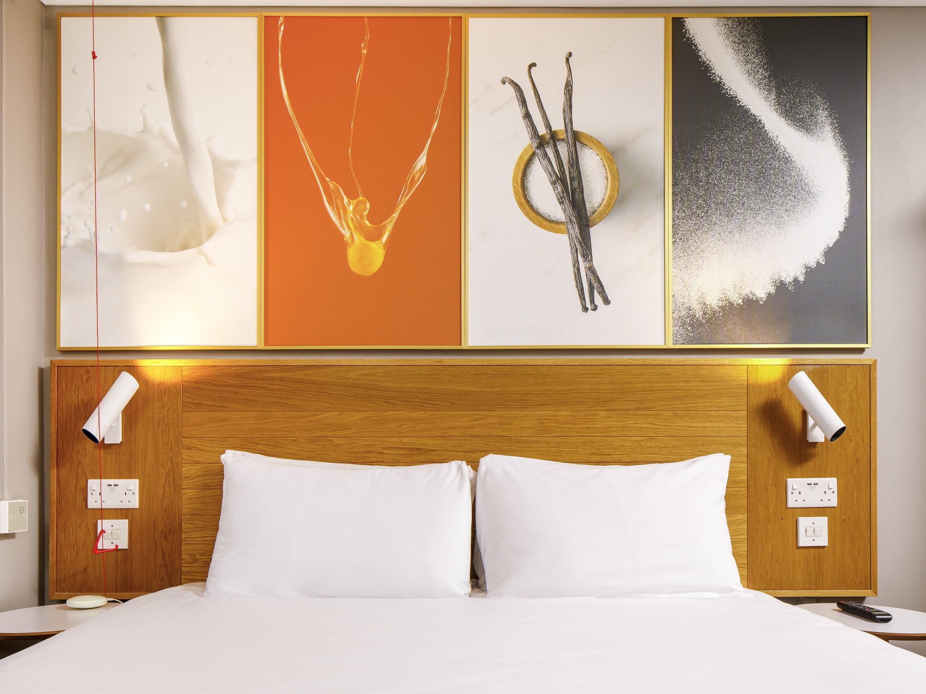 A hotel room with a wooden headboard, two bedside lamps, a bed with white bedding, and a wall with four framed artworks.