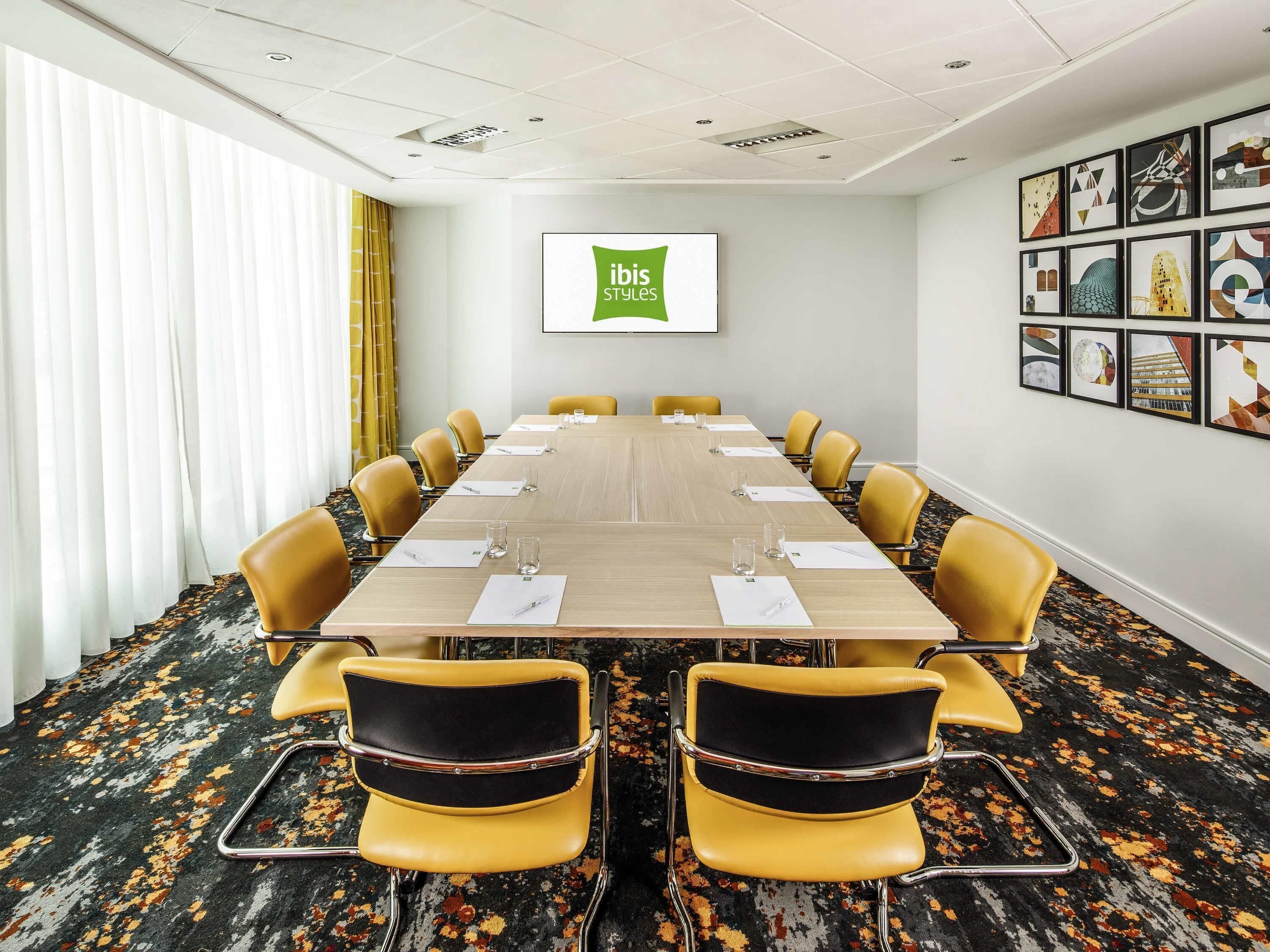 A conference room with a long table, yellow chairs, and a 'ibis Styles' sign on the wall.