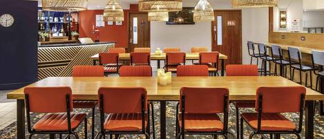 A hotel dining area with a long wooden table surrounded by orange chairs, overhead wicker chandeliers, and a carpeted floor.