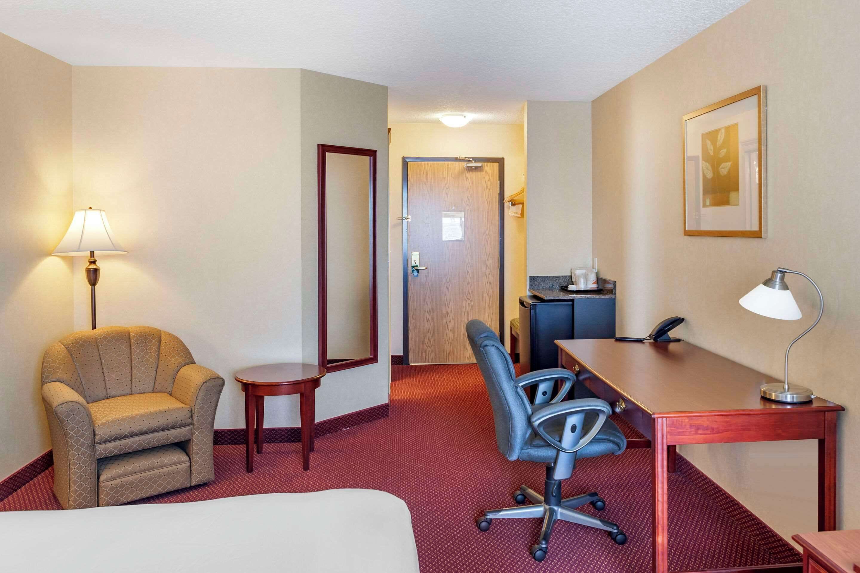 Ett hotellrum med en säng, ett skrivbord, en stol, en lampa, ett litet bord och en dörr som leder till ett annat rum.