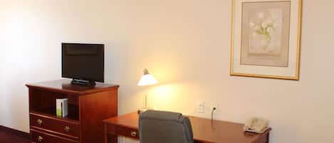 A hotel room with a wooden desk, a chair, a television on a cabinet, a lamp, a telephone, and a framed picture on the wall.