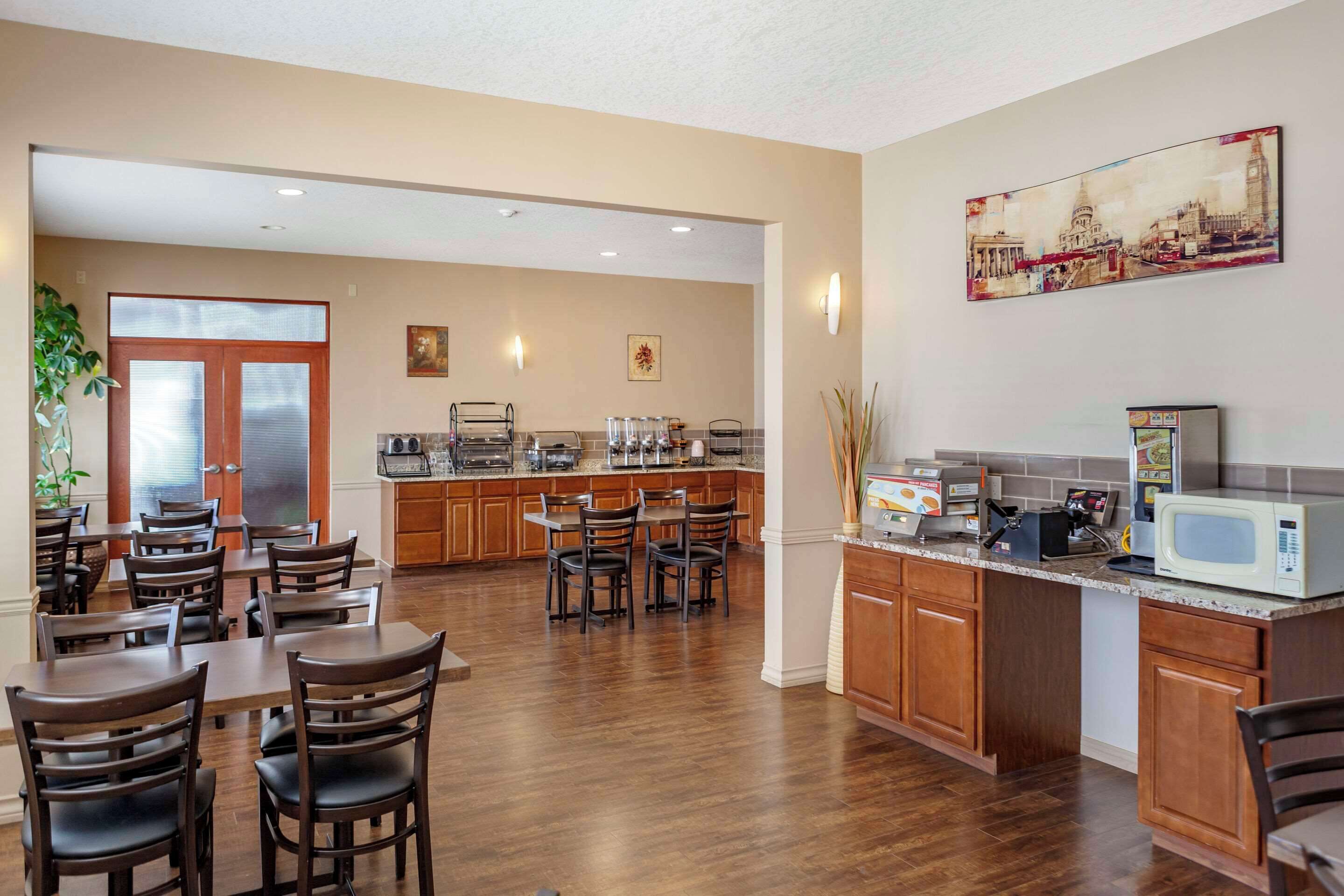 A hotel kitchen with wooden cabinets, a dining area with tables and chairs, a microwave, and a painting on the wall.