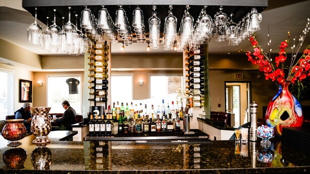 A bar area with a wine and liquor display, a decorative vase with red flowers, and a man sitting at a table. - Quality Inn & Suites Edmonton International Airport (Leduc)