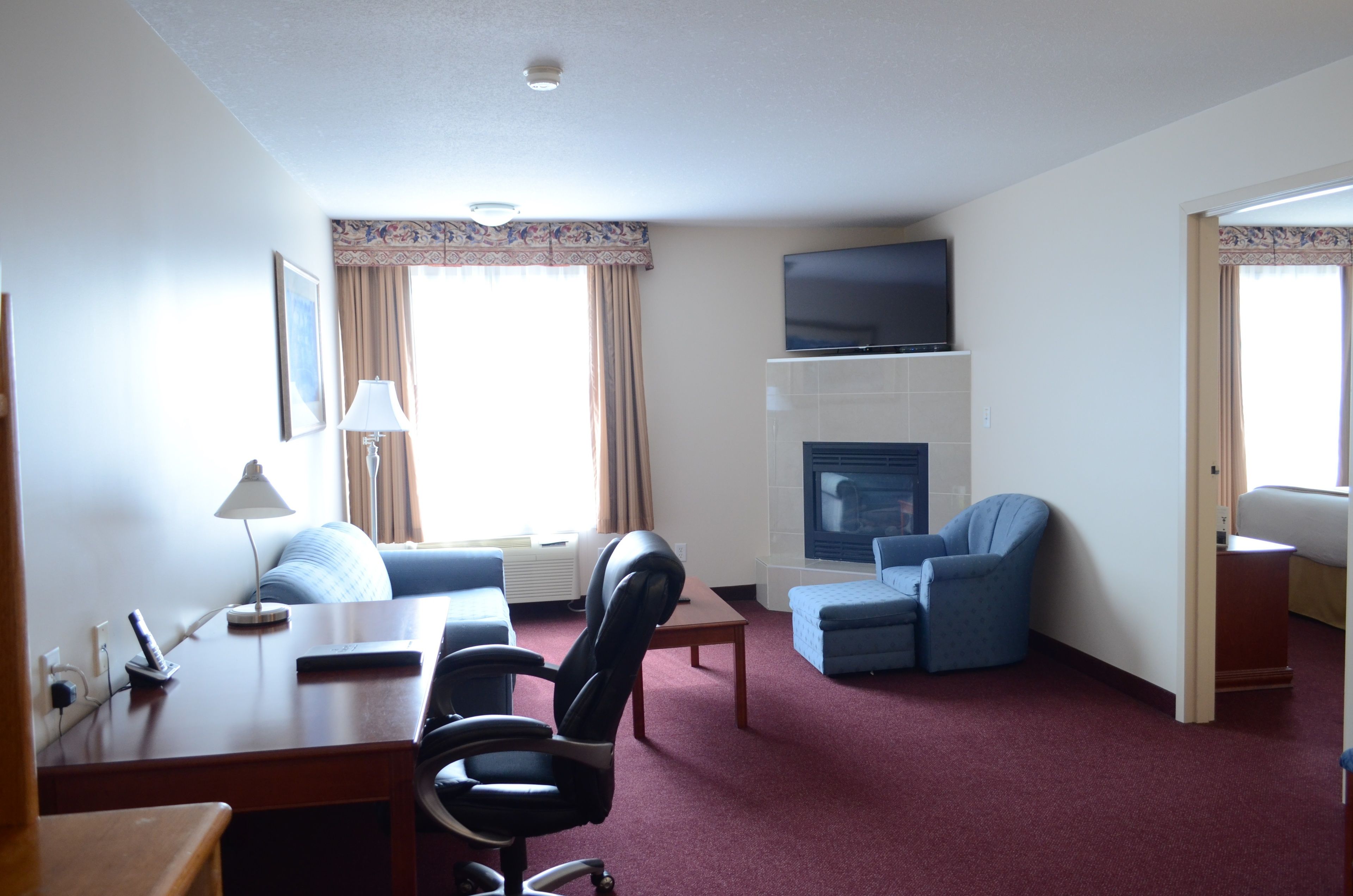 Habitación de hotel con sala de estar, televisor de pantalla plana sobre la chimenea, escritorio con lámpara y sillón azul.