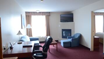A hotel room with a living area, a flat-screen TV mounted above a fireplace, a desk with a lamp, and a blue armchair.