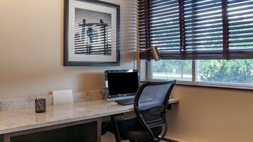 Un espacio de trabajo con una computadora de escritorio, una silla, un cuadro enmarcado y una ventana con persianas.