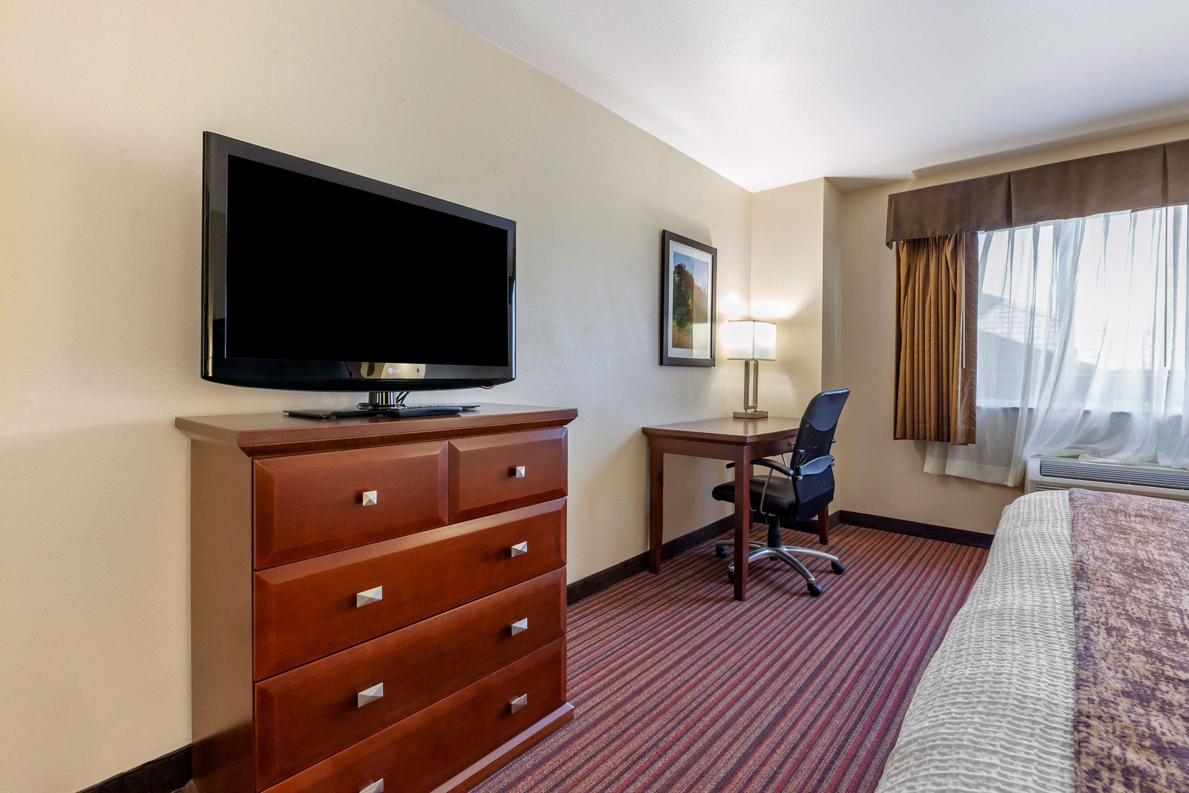 A hotel room with a flat-screen TV, a desk with a chair, a bed, and a window with curtains.