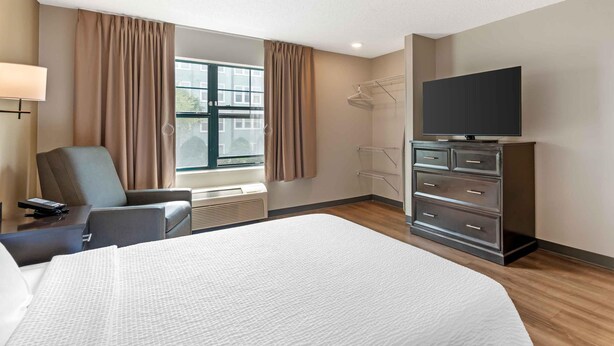 A hotel room with a bed, a desk with a lamp, a chair, a TV on a dresser, and a window with curtains.