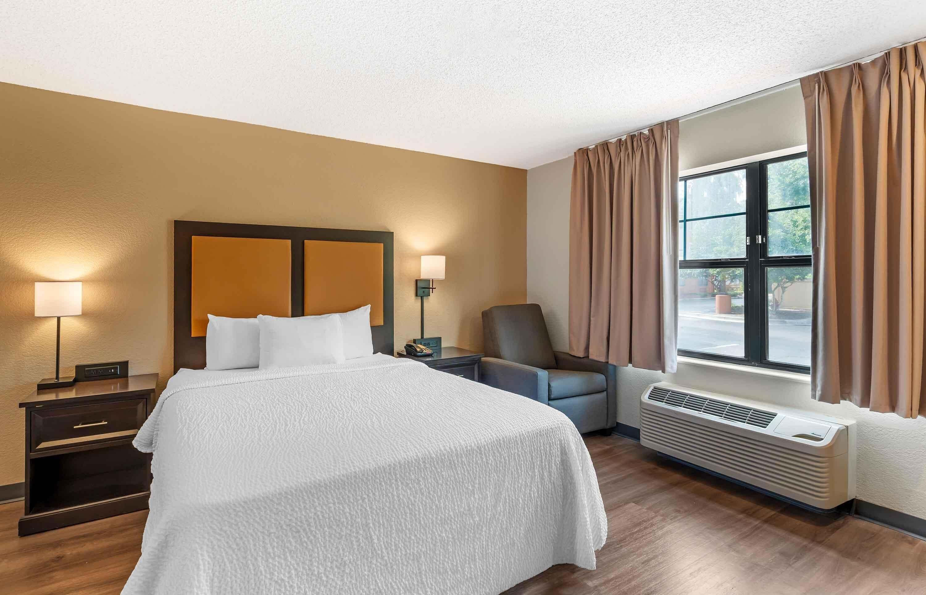 A hotel room with a bed, bedside lamps, a chair, a window with curtains, and a wall-mounted air conditioner.